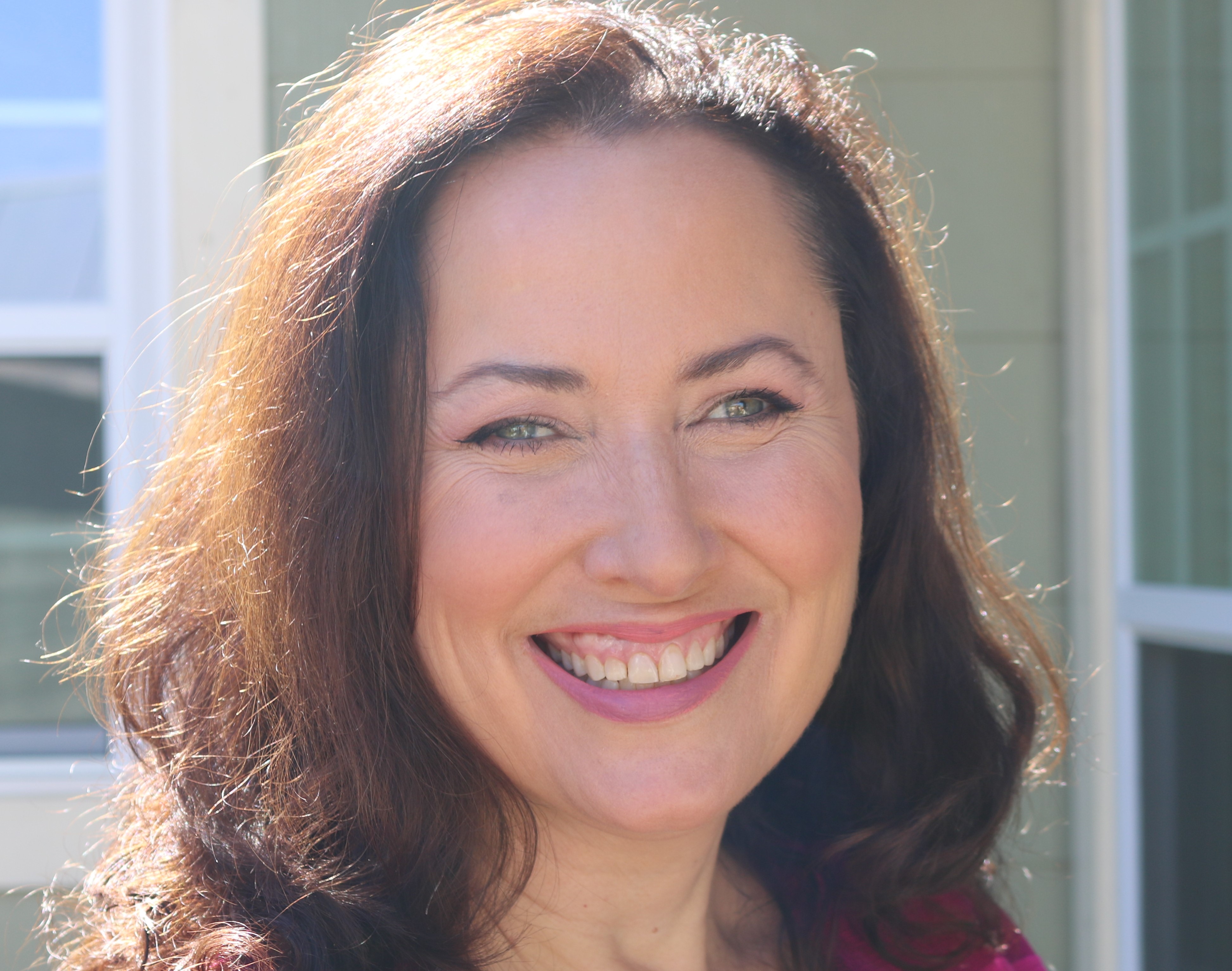 A middle aged brunette woman with shoulder length wavy hair smiles widely at the camera. She wears natural make up and has green eyes. The lighting is soft and she is pictured outdoors. Insertion at beginning of text.